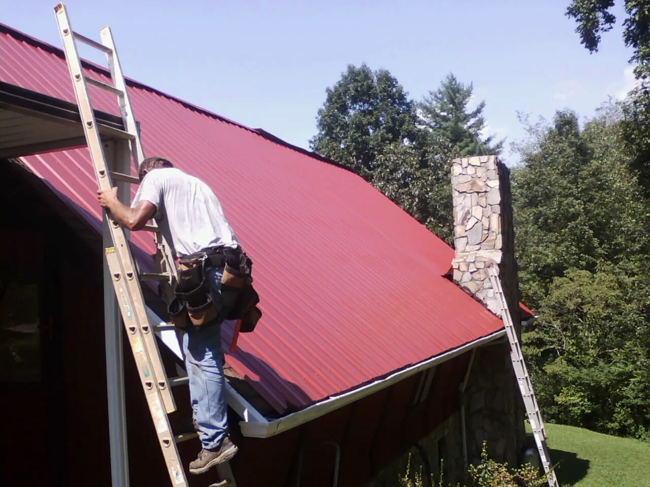 Roofer on a ladder beside a red metal roof and stone chimney — Soffit & Fascia Repair by Roofing Brothers in Pine City